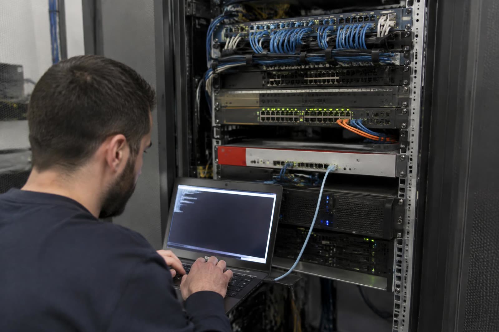 Técnico configurando switches y un firewall montados en rack desde una laptop conectada por consola.